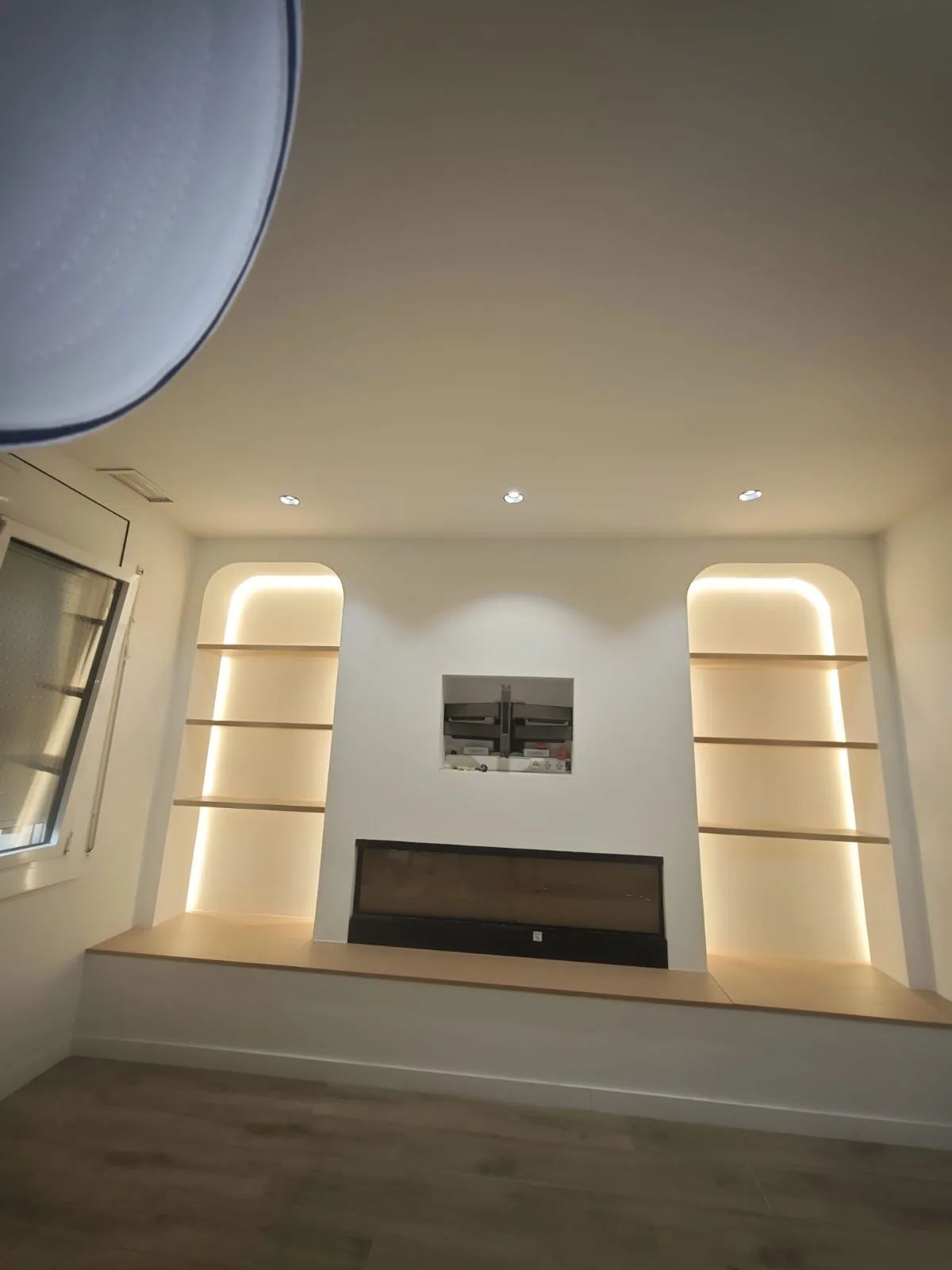 Salón con estanterías de madera empotradas en hornacinas con luz LED y banco corrido bajo la chimenea