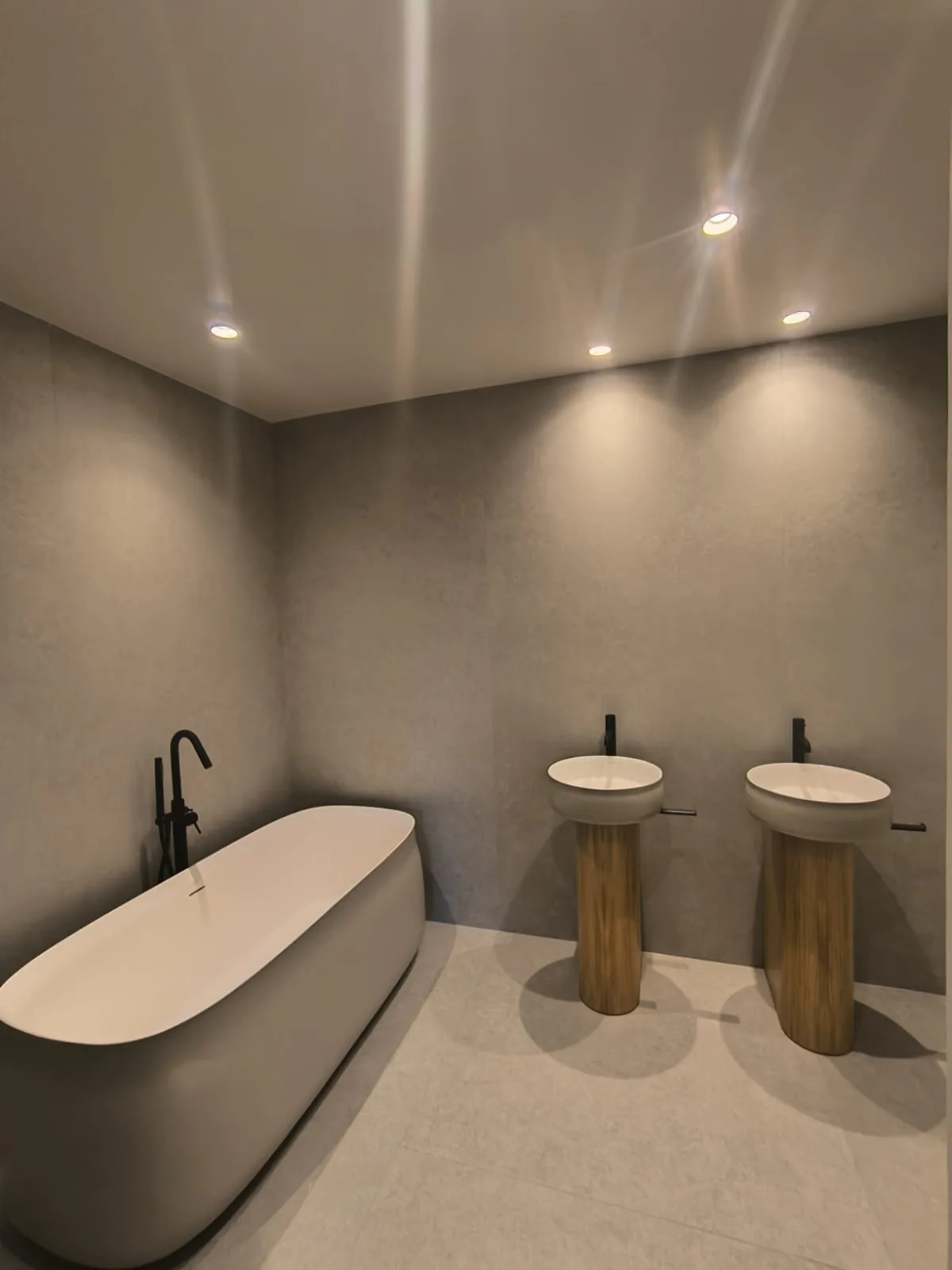 Baño amplio con bañera exenta, dos lavabos sobre pie de madera y paredes de microcemento gris