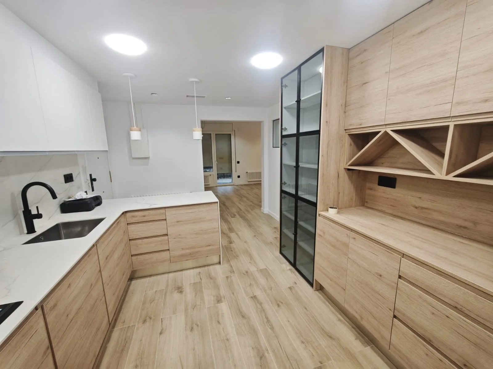 Cocina en L con encimera blanca, muebles de madera clara, fregadero negro y mueble con vinoteca