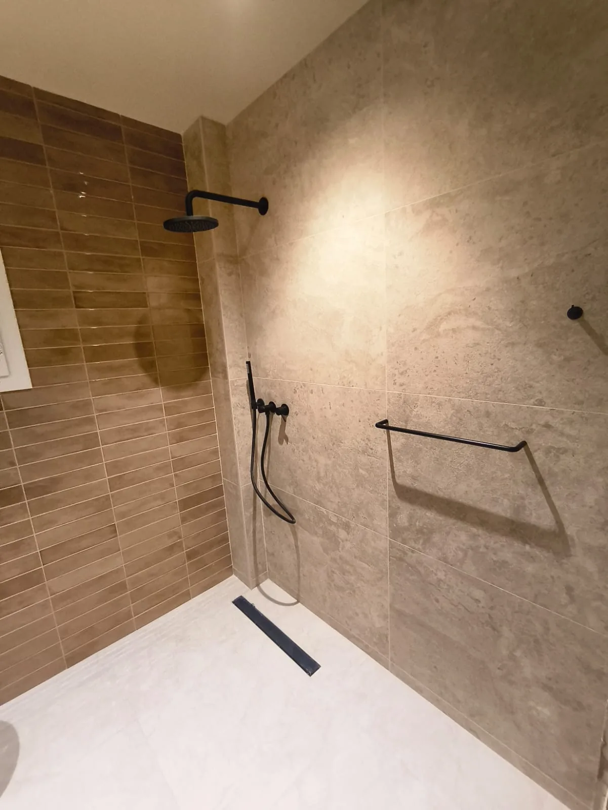 Ducha a ras de suelo con desagüe lineal, grifería negra y revestimiento de gres en tonos arena