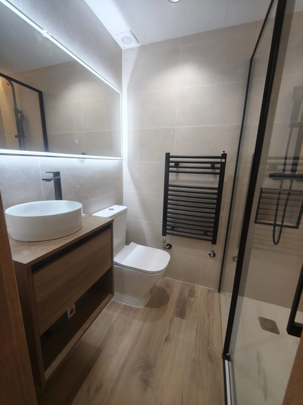 Baño con mueble de madera suspendido, lavabo sobre encimera, espejo LED, toallero negro y mampara de ducha