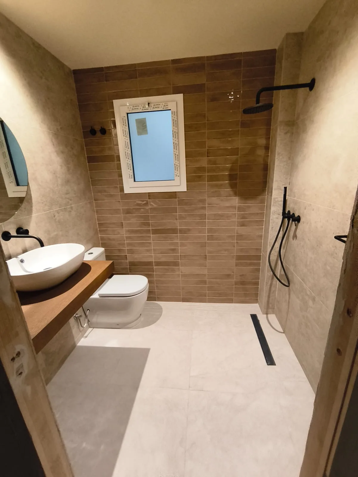 Baño reformado con ducha de obra, azulejos en tonos arena y madera, lavabo sobre encimera de madera y grifería negra